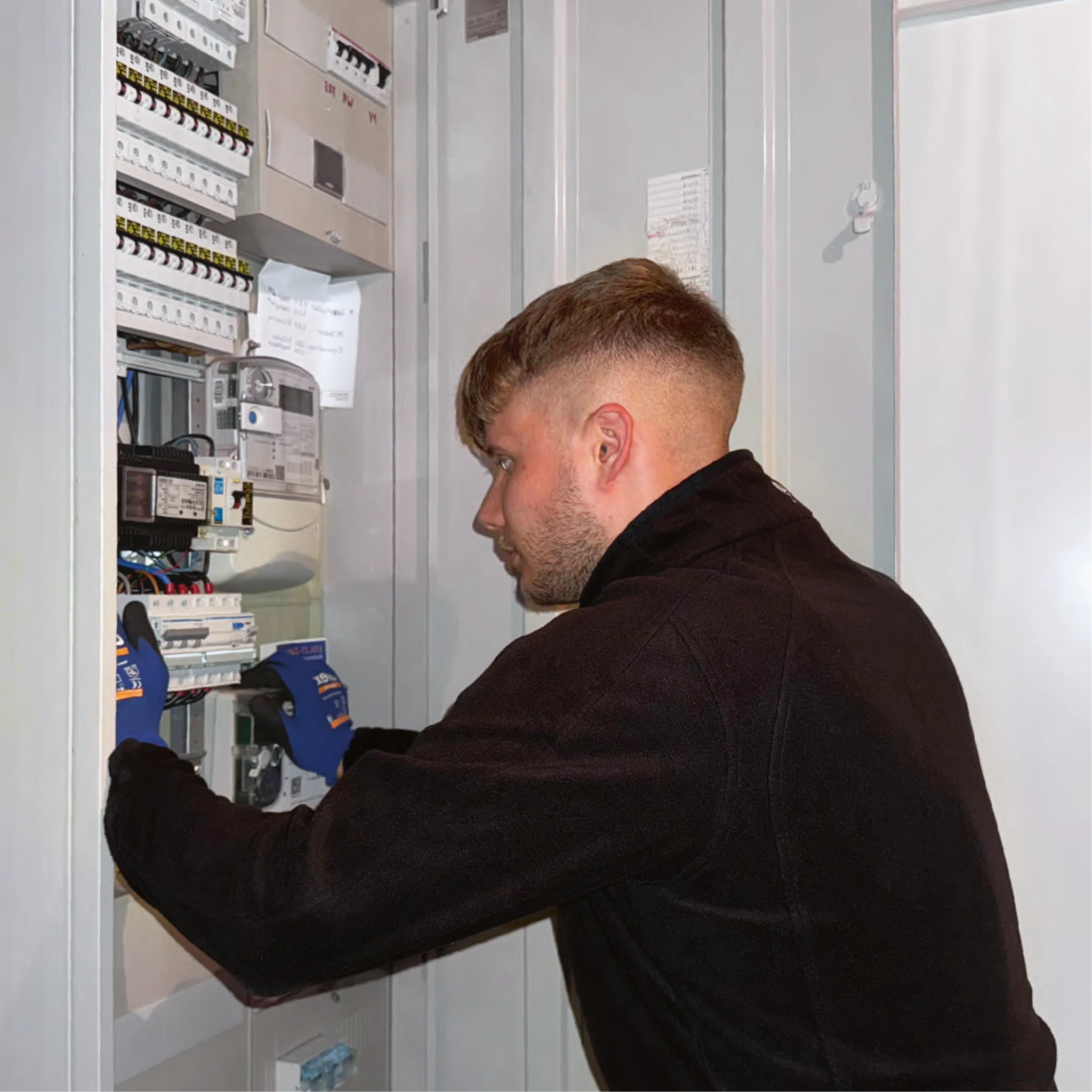 Elektriker in schwarzer Arbeitsjacke verdrahtet mit blauen Handschuhen einen geöffneten Zählerschrank